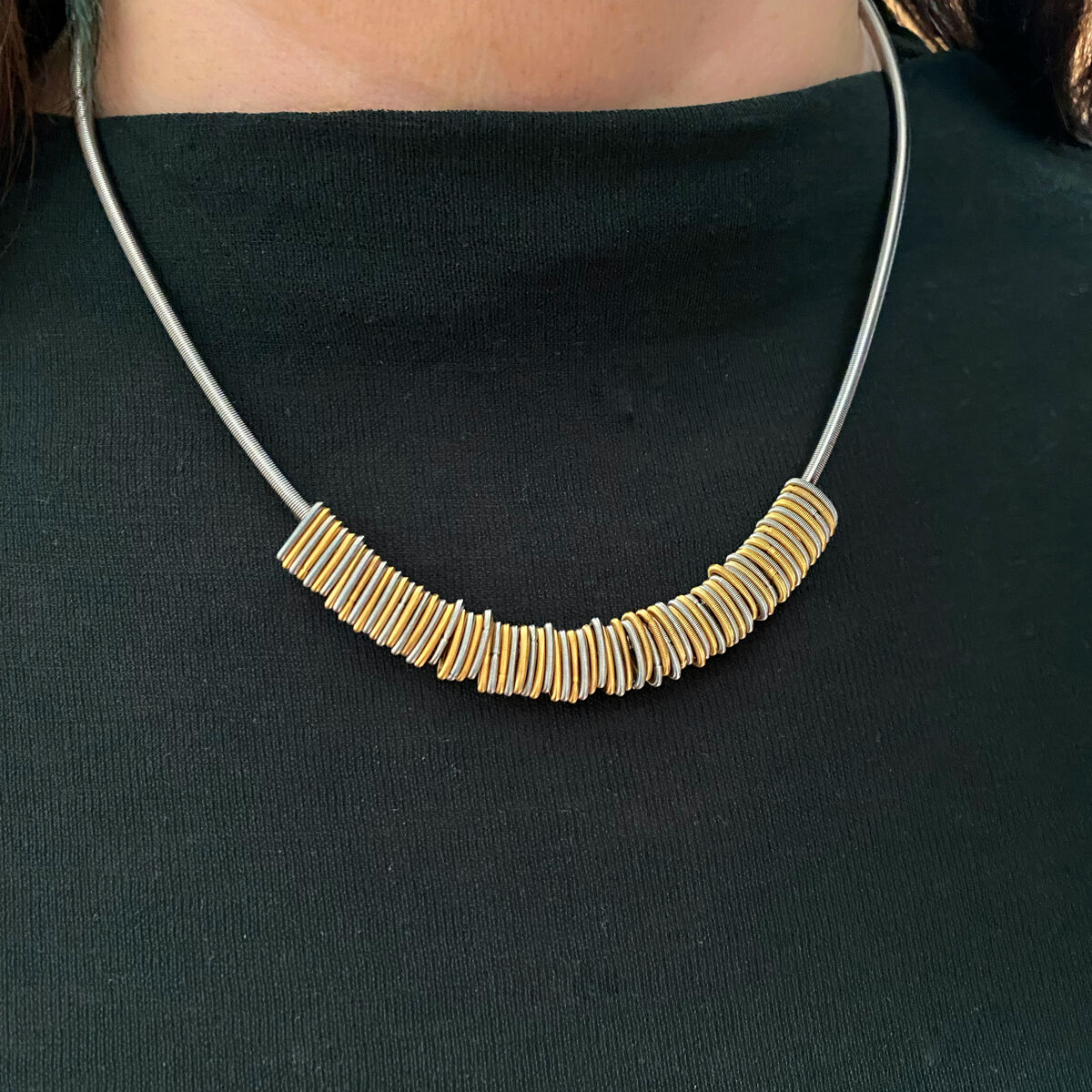 Nahaufnahme einer eleganten Halskette aus der ZEIT-Sonderedition. Die Kette besteht aus einem schmalen metallischen Band, an dem zahlreiche goldene, rechteckige Elemente aufgereiht sind. Das Schmuckstück wird auf einem schwarzen Oberteil präsentiert.
