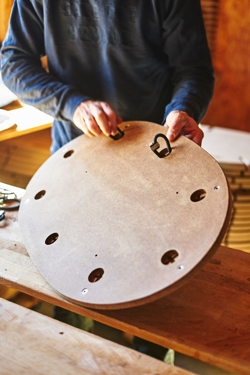 Eine Person hält ein rundes Sensoboard aus Sperrholz mit einem Cutout-Design. Das Balancebrett hat mehrere Löcher und zwei Griffe. Die Person trägt einen dunkelblauen Pullover. Im Hintergrund ist eine Holzstruktur zu erkennen.
