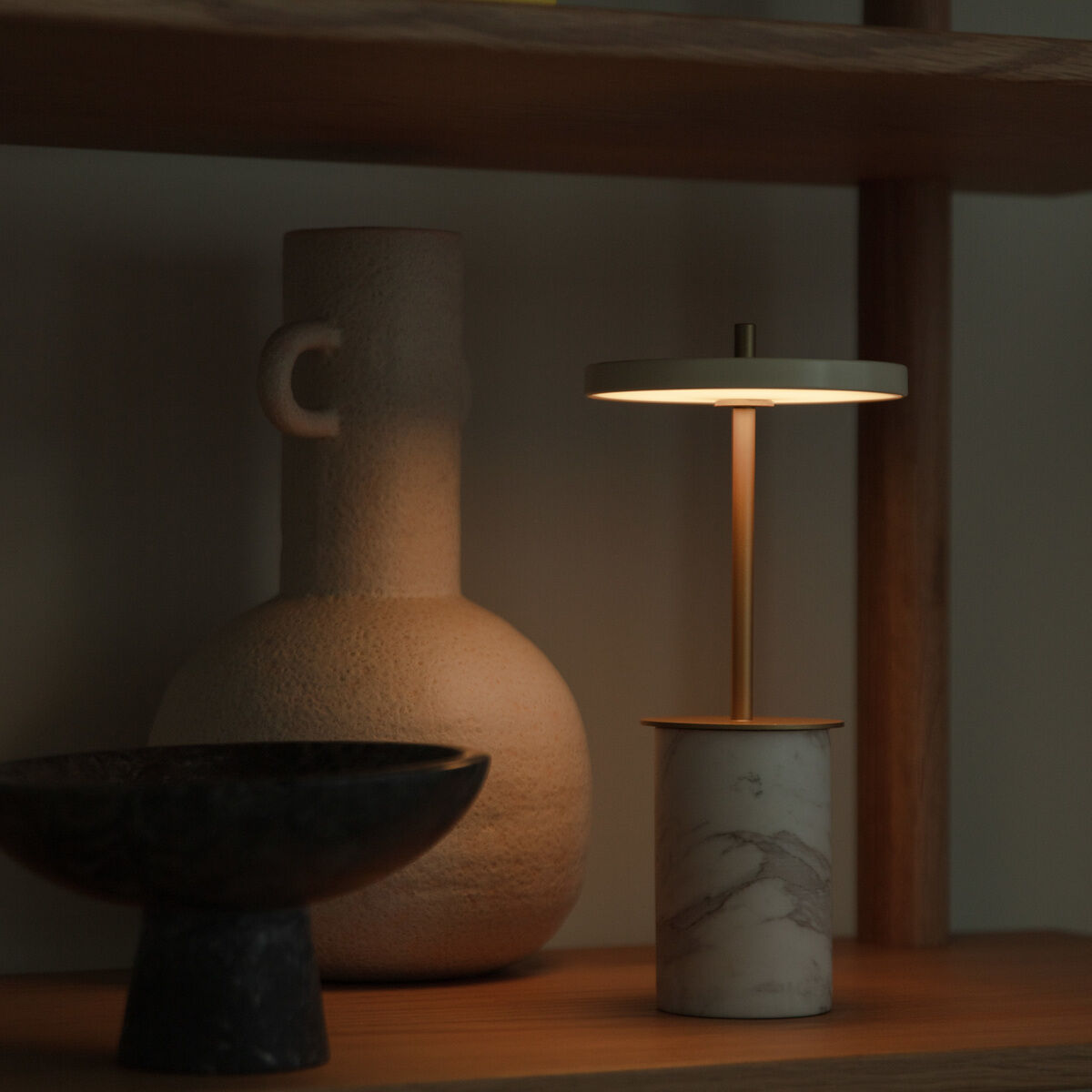 Auf einem Holzregal steht eine elegante, kabellose Tischlampe Asteria Move Mini mit Marmorfuß und goldenem Stab. Die Lampe leuchtet warm und steht neben einer strukturierten Vase und einer Schale, gemeinsam mit Büchern. Das Bild vermittelt ein modernes, minimalistisches Wohnambiente.