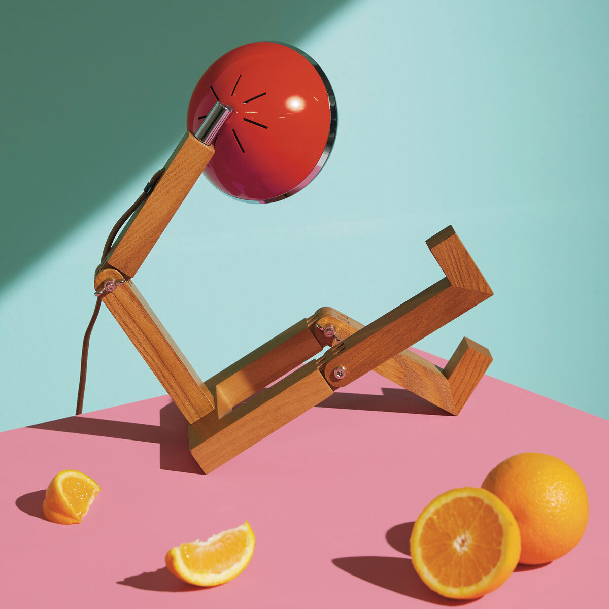 Eine Tischlampe namens 'Mr. Wattson' mit einem kreativen Design, das an eine sitzende Figur erinnert. Die Lampe hat einen verstellbaren Holzrahmen und einen orangefarbenen Lampenschirm. Sie steht auf einem pinkfarbenen Hintergrund, vor einer mintgrünen Wand mit Orangenstücken als Dekoration.