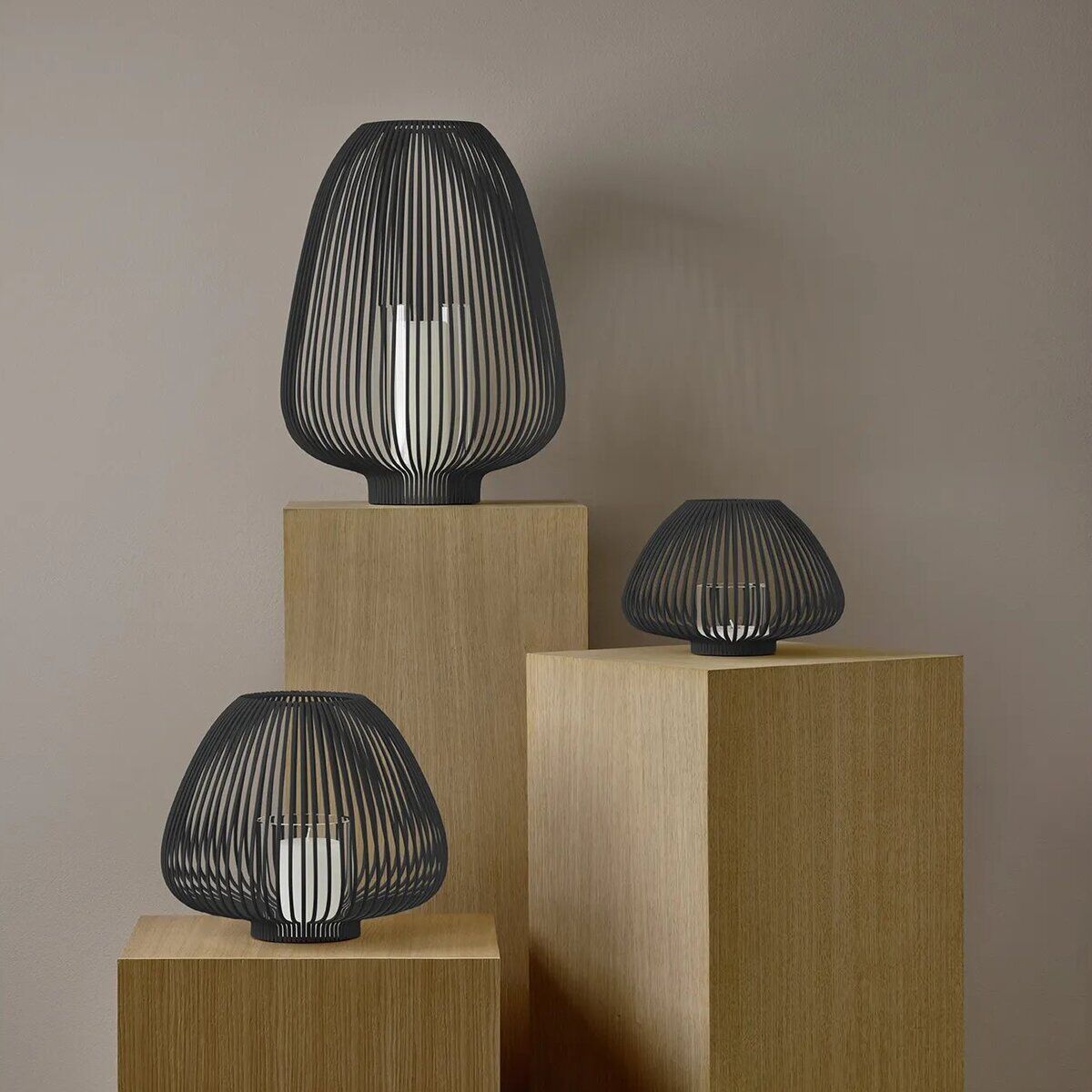 Drei moderne schwarze Metall-Laternen in unterschiedlicher Größe stehen auf naturfarbenen Holzsockeln vor einer beigen Wand. Die Laternen sind leer oder mit Kerzen bestückt und schaffen eine minimalistische, stilvolle Atmosphäre für den Innen- oder Außenbereich.