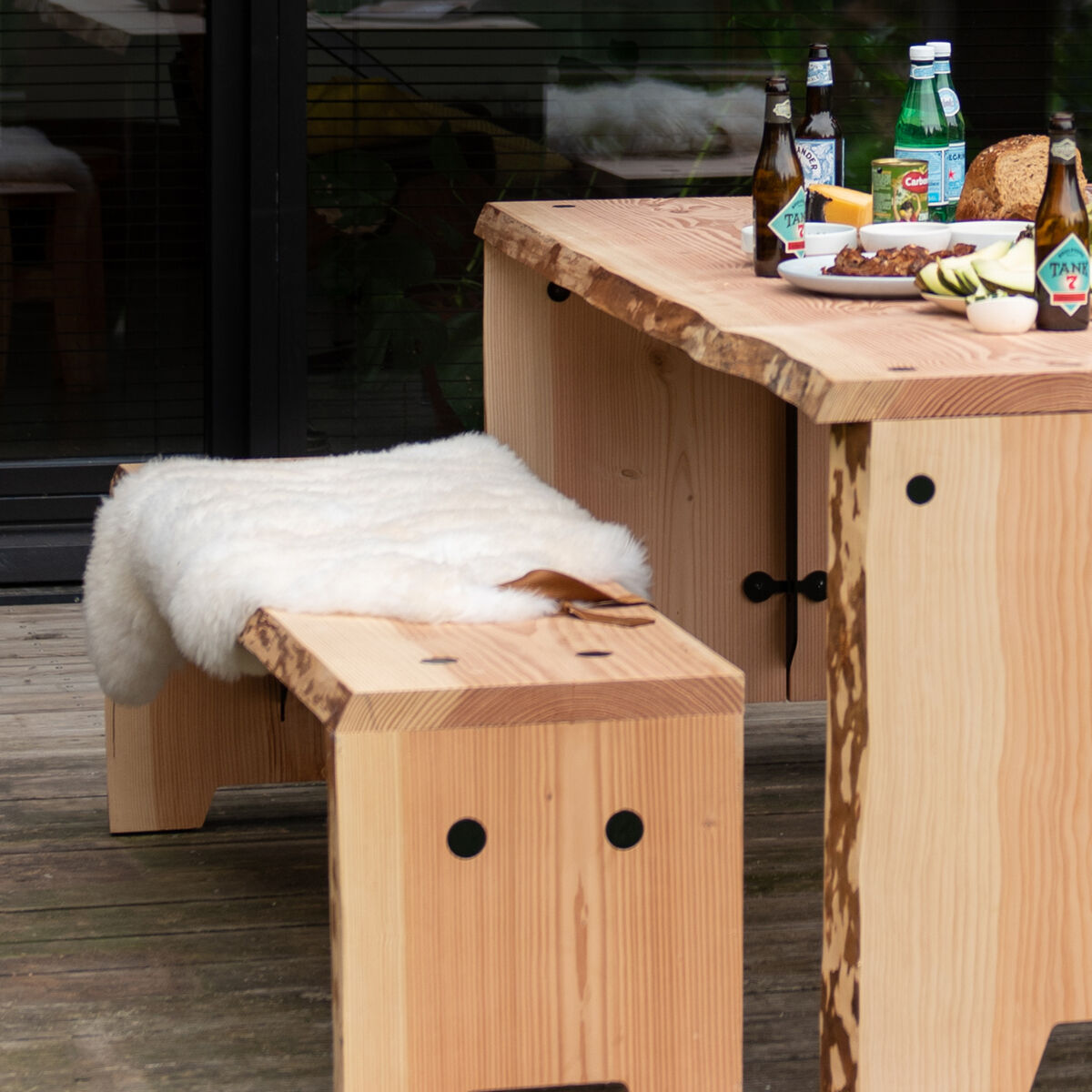 Ein naturweißes Schafsfell mit Lederschlaufe liegt dekorativ auf einer hellen Holzbank, die neben einem Holztisch auf einer Terrasse steht. Auf dem Tisch befinden sich Speisen, Getränke und Geschirr. Die Szene vermittelt eine gemütliche, naturnahe Atmosphäre im Außenbereich.