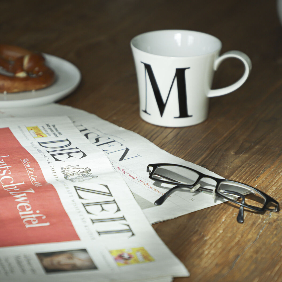 Eine weiße Frühstückstasse mit dem Buchstaben 'M' darauf steht auf einem Holztisch neben einer Zeitung 'Die Zeit', einer Brille und einer Brezel.