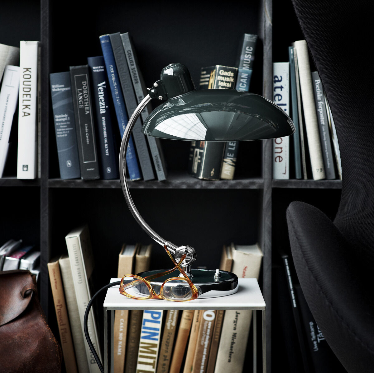 Ein modernes, schwarzes Tischlampe-Modell namens 'Kaiser Idell' steht auf einem weißen Schreibtisch vor einem Wandregal voller Bücher. Die Lampe hat ein glänzendes Metallgestell und geschwungene Linien. Daneben befindet sich eine Brille sowie eine schwarze Stuhllehne am rechten Bildrand.