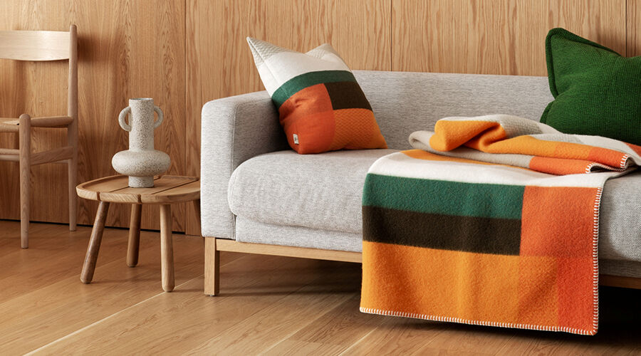 Hellgraues Sofa mit buntem Kissen und Decke in Orange, Grün und Braun vor einer Holzpaneelwand, daneben ein kleiner Holztisch mit dekorativer Vase und ein Holzstuhl