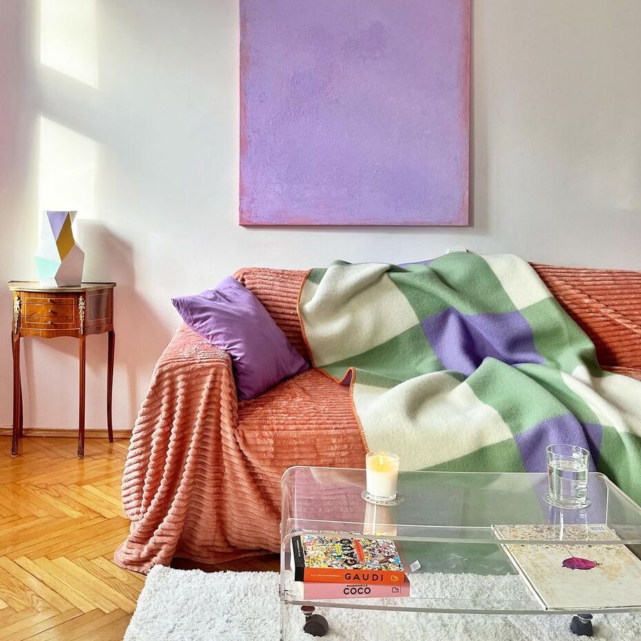 Ein Wohnzimmer mit einem Sofa, das mit einer Decke in grün-lila Karos bedeckt ist. Daneben ein runder Holztisch und ein durchsichtiges Couchtisch mit Büchern. Ein purpurfarbener Vorhang und ein pastellrosa Bild an der Wand vervollständigen die gemütliche Atmosphäre.