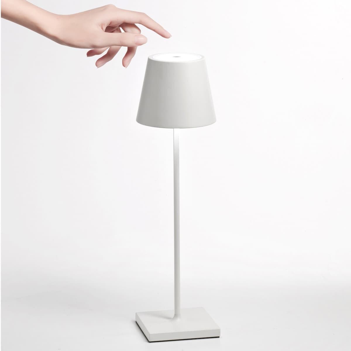 Eine schlichte, weiße LED-Tischlampe mit dem Namen »Poldina Pro«, die auf einem glatten Untergrund steht. Eine Hand über der Lampe deutet auf deren Bedienbarkeit durch Berührung hin. Die Lampe stellt ein minimalistisches Design und eine stimmungsvolle Beleuchtung dar.