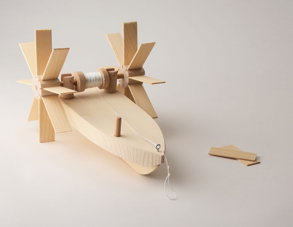 Ein Bausatz aus Holz zum Experimentieren mit dem Namen 'Forelle'. Der Bausatz besteht aus mehreren Holzteilen, darunter eine Basis in Fischform und zwei Propellern. Der Hintergrund ist schlicht und einfarbig, wodurch das Holzspielzeug im Mittelpunkt steht.