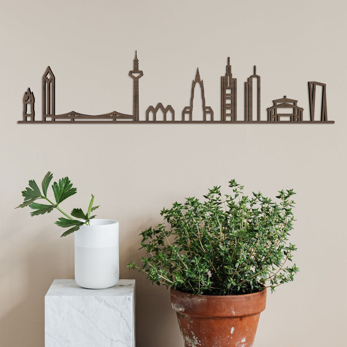 Dekorative Holzkunst-Skyline von Frankfurt a.M. an einer beige gestrichenen Wand montiert. Darunter stehen ein weißer Pflanzkübel mit grüner Pflanze auf einem weißen Podest und ein größerer Terrakottatopf mit einer weiteren Pflanze. Moderne und stilvolle Wanddekoration geeignet für Wohnungen und Büros.