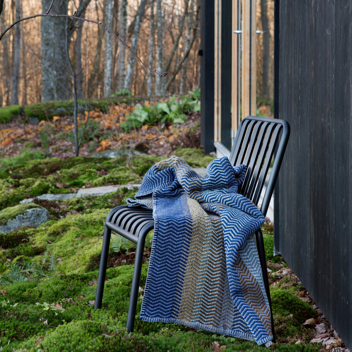 Eine Lammwolldecke in verschiedenen Blau- und Brauntönen liegt über einem schwarzen Stuhl, der im Wald neben einem modernen Holzhaus steht. Moosbedeckter Boden und Bäume im Hintergrund.