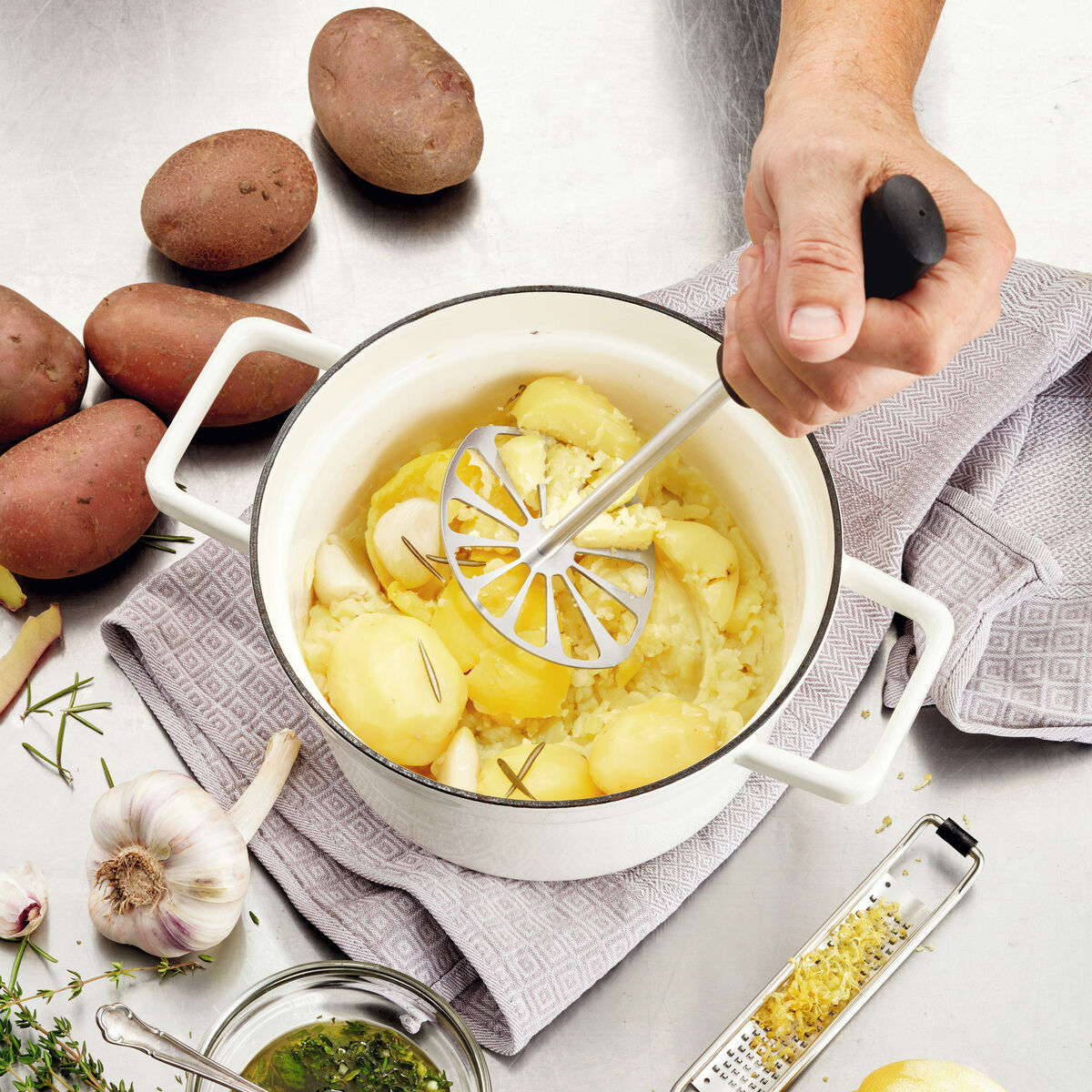 Ein eleganter Kartoffelstampfer »Soul« von triangle mit Griff aus Kirschholz und Edelstahl-Stampfplatte wird in einem Topf mit gekochten Kartoffeln genutzt. Umgeben von roten Kartoffeln, einer Knoblauchknolle, Kräutern, Zitronenabrieb und Küchenutensilien auf einem hellen Tuch.