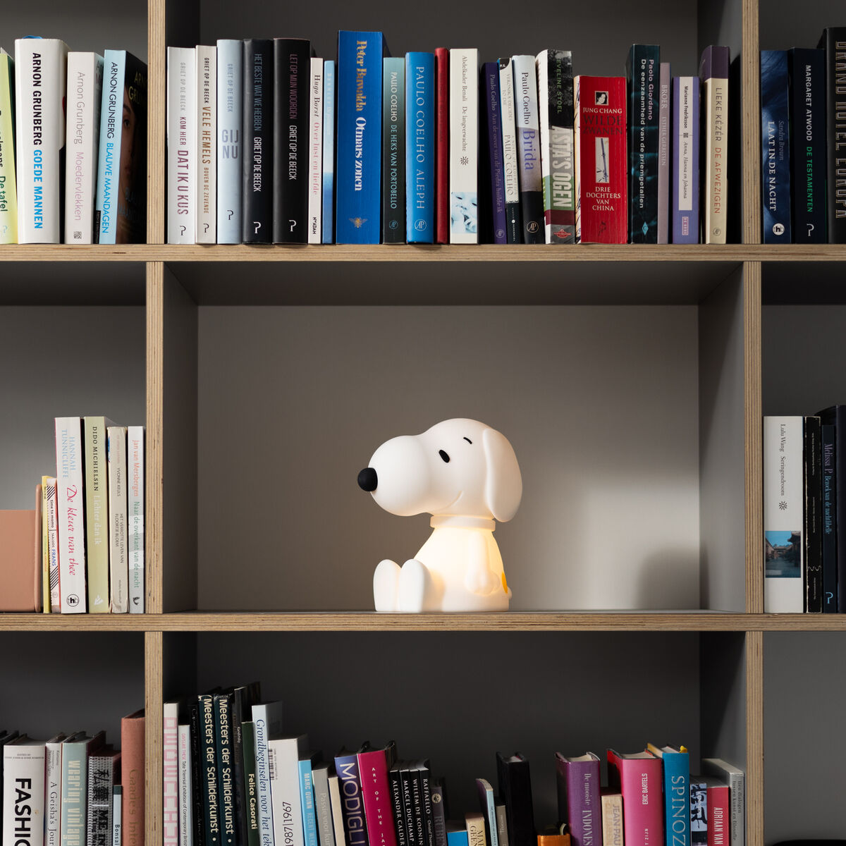 Ein Nachtlicht in Form der Comicfigur Snoopy sitzt in einem Bücherregal mit verschiedenen Büchern. Das Licht des Snoopy-Nachtlichts erleuchtet die Umgebung dezent und schafft eine gemütliche Atmosphäre.