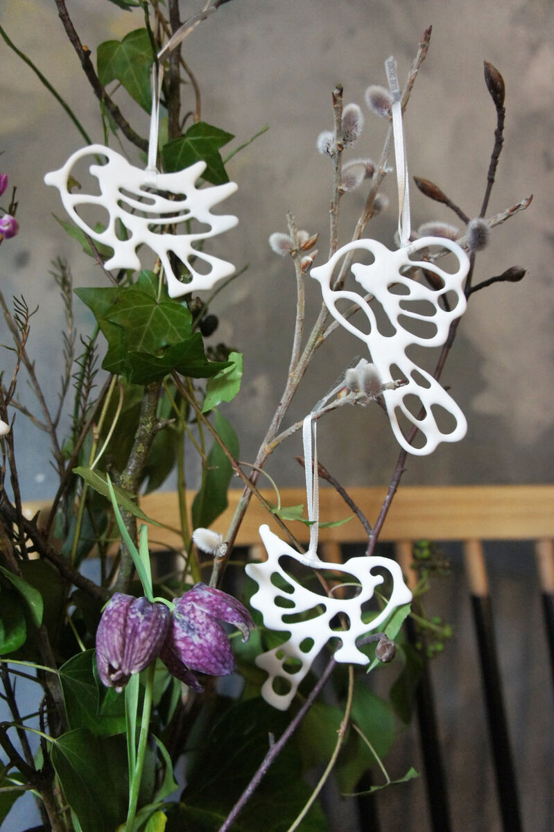 Sechs weiße Porzellanvögel hängen an Zweigen, die mit grünen Blättern und violetten Blüten dekoriert sind. Die Vögel haben ein filigranes, ausgeschnittenes Design und sind als dekorative Anhänger angebracht.