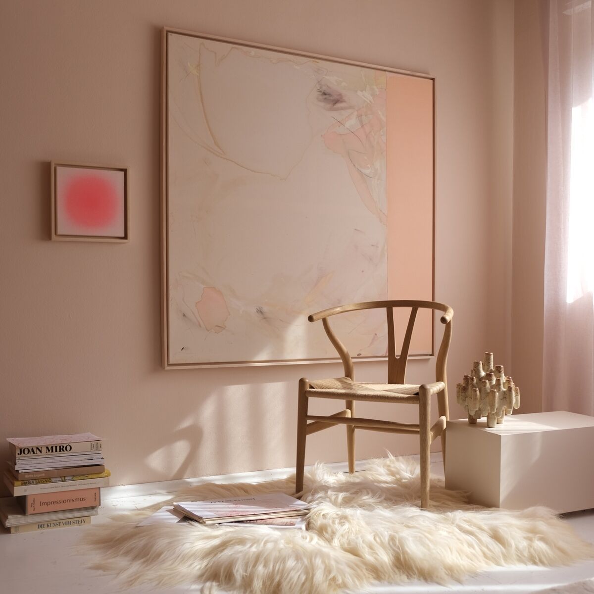 Im Vordergrund steht ein moderner, abstrakter Kunstdruck in Rosa- und Beigetönen mit Holzrahmen an einer pastellrosa Wand. Eine minimalistische Einrichtung mit hellbraunem Sessel, Teppich aus Kunstfell und indirektem Tageslicht schafft eine warme, einladende Atmosphäre. Links sind weitere Kunstwerke sowie Designbücher sichtbar.