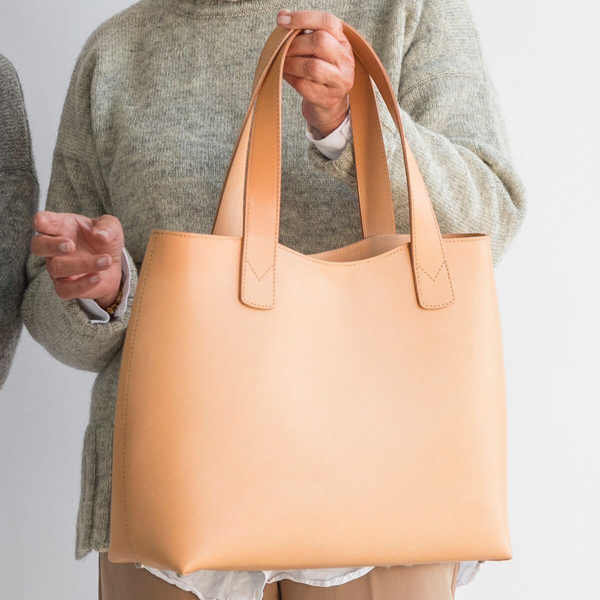 Eine große Tasche aus hellem Kernleder, getragen von einer Person in einem grauen Pullover. Die Tasche hat eine schlichte, minimalistische Form mit zwei Griffen. Sie wirkt hochwertig und zeitlos.
