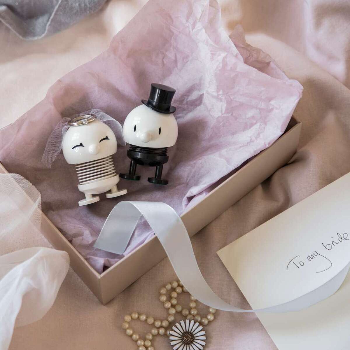 Zwei Hoptimist-Figuren als Braut und Bräutigam sitzen in einer Geschenkbox auf rosa Papier. Die Braut trägt einen Schleier, der Bräutigam einen Zylinder. Daneben liegen eine weiße Schleife, Perlen, Blütenbrosche und ein Umschlag mit der Aufschrift 'To my bride'. Freundlicher, heller Hintergrund.