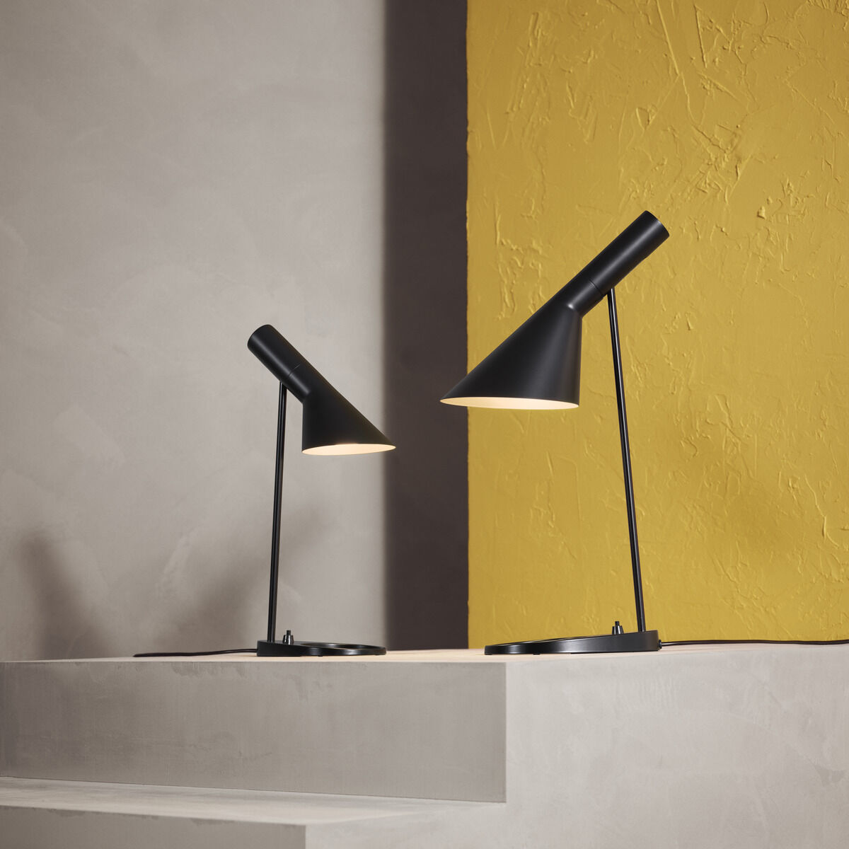 Zwei schwarze Tischlampen des Modells AJ Table Mini stehen auf gestaffelten weißen Plattformen vor einer zweifarbigen Wand in gelb und grau.