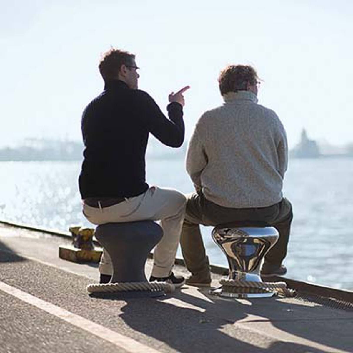 Zwei Personen sitzen auf Hamburger Hockern mit Segeltau an einem Kai mit Blick aufs Wasser. Links trägt eine Person eine dunkle Jacke, rechts eine graue Jacke. Ein heller Tag mit klarem Himmel.