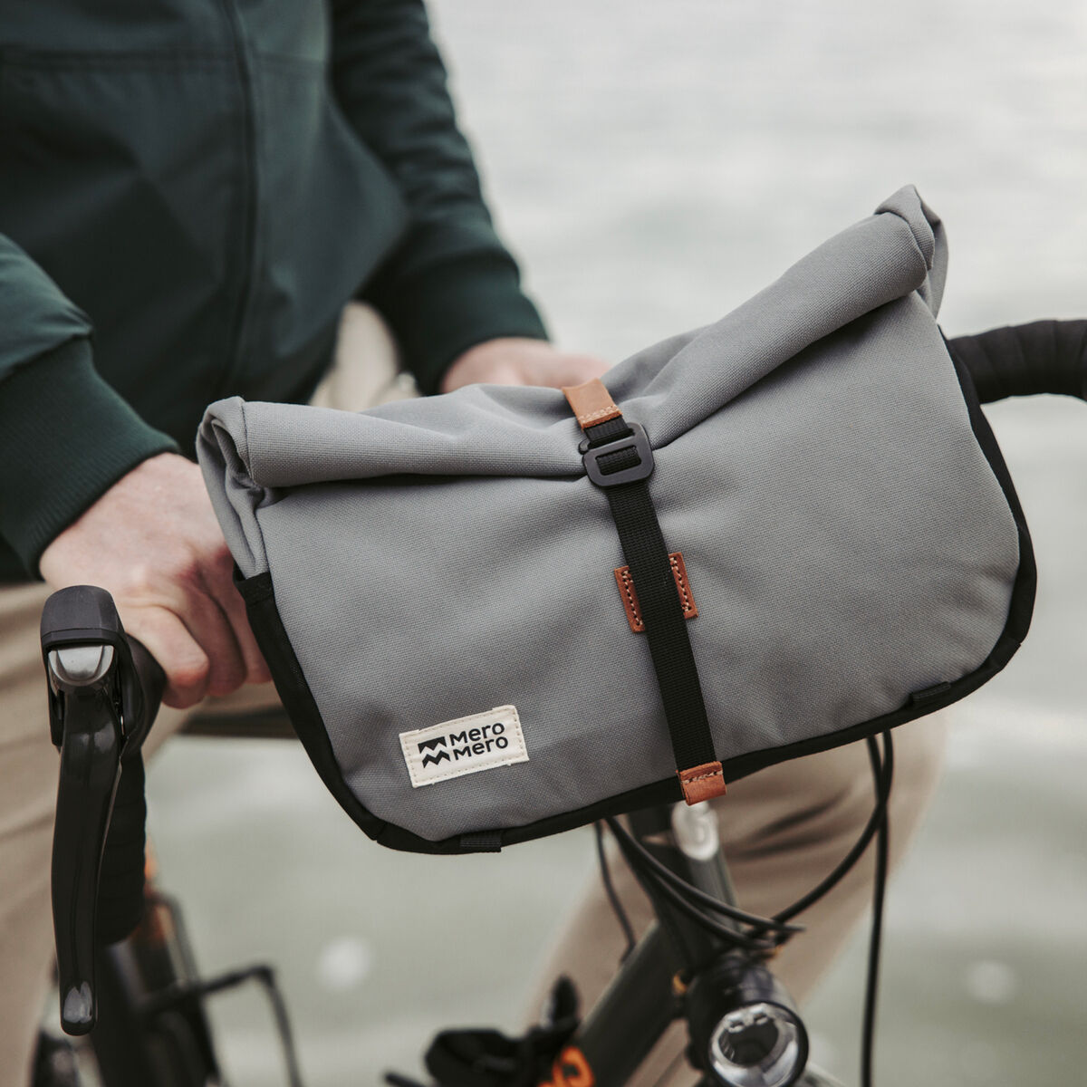 Nahaufnahme einer grauen Fahrradtasche, befestigt an einem Fahrradlenker. Die Tasche hat einen schwarzen Riemen und braune Lederdetails und wird von einer Person in einem grünen Pullover und beigen Hosen gehalten.