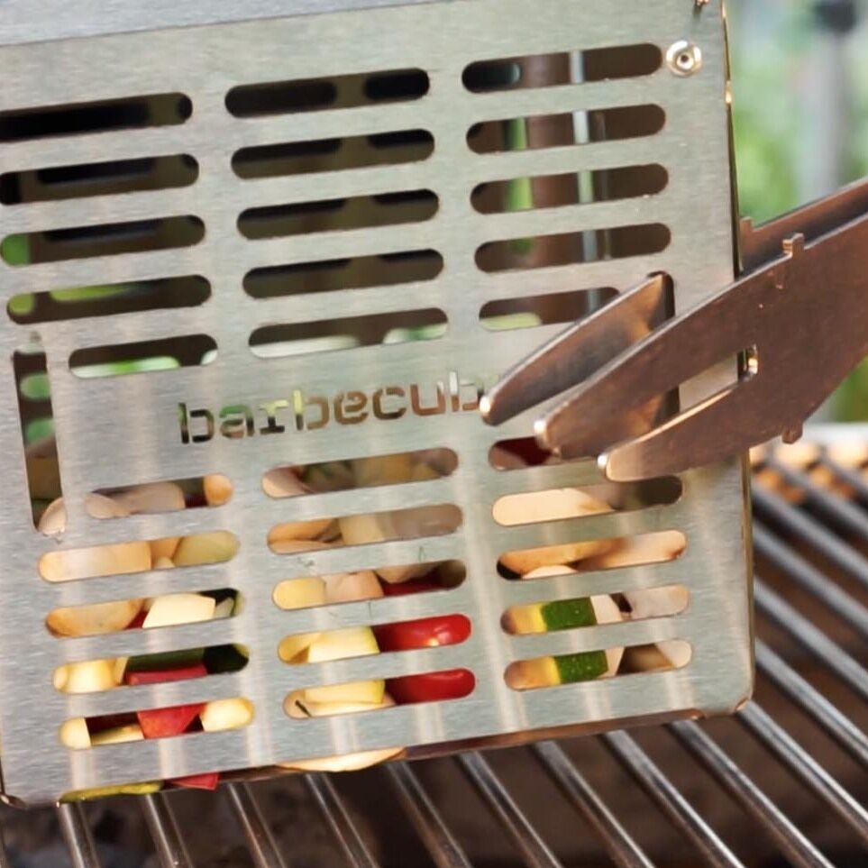 Eine Nahaufnahme eines Grillwürfels aus Edelstahl, bezeichnet als »The Cube«, der auf einem Grillrost platziert ist. Eine Hand hält eine Grillzange und bewegt den Grillwürfel. Im Inneren des Würfels sind bunte Gemüse wie rote und grüne Paprika zu sehen.