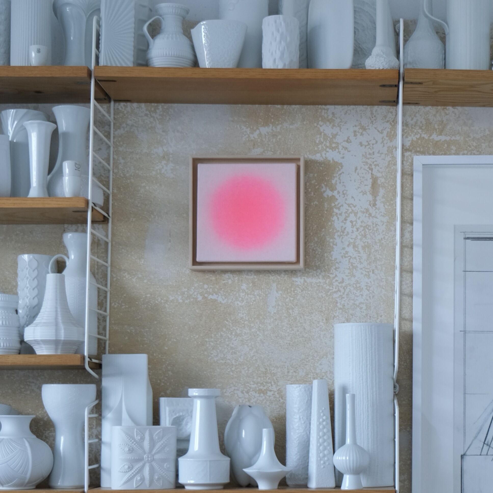 Quadratisches Kunstwerk in kräftigem Pink, mittig an einer beige strukturierten Wand. Es ist in hellem Holz gerahmt. Rundherum stehen auf Holzregalen zahlreiche weiße Vasen und Keramiken, das Gesamtbild wirkt modern und minimalistisch.