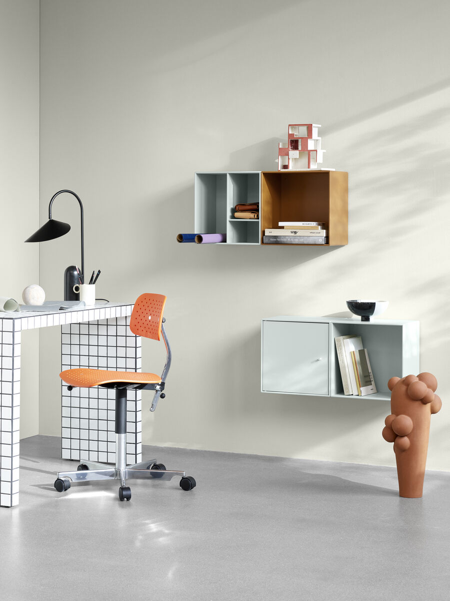 Ein moderner Arbeitsplatz mit einem weiß gefliesten Schreibtisch, einem oranges Bürostuhl und flachen Wandregalen in verschiedenen Farben. Auf dem Schreibtisch befinden sich mehrere Dekorationsgegenstände und neben dem Tisch steht eine skulpturale Vase.