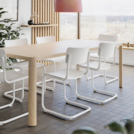 Großer, heller Essbereich mit sechs weißen Thonet Freischwingern »S 43 Black« um einen rechteckigen Holztisch auf grauen Fliesen. Über dem Tisch hängt eine rote Lampe. Große Fenster bieten Blick in die Landschaft, Pflanzen ergänzen das moderne, minimalistische Ambiente.