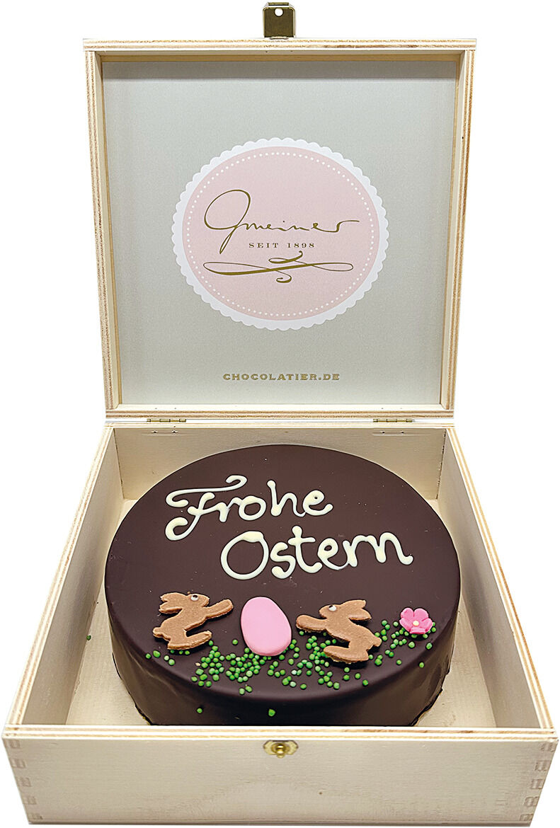 Eine Sachertorte in einer hellen Holzbox mit der Aufschrift 'Frohe Ostern' und dekoriert mit Osterhasen und grünen und rosa Ornamenten.