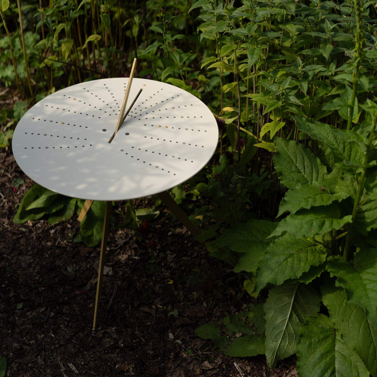 Eine Sonnenuhr steht in einem üppigen Garten, umgeben von grünen Pflanzen und rotem Laub. Die Sonnenuhr ist aus Metall gefertigt und zeigt zeitliche Markierungen.