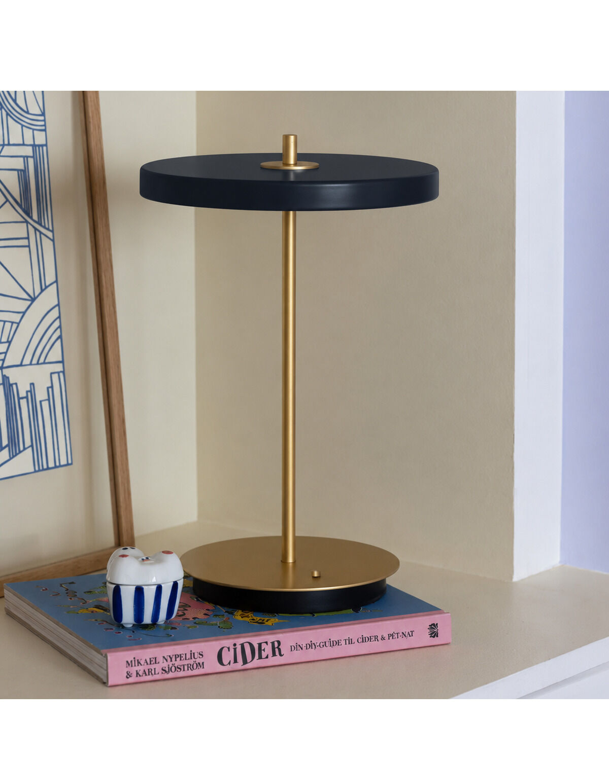 Die ZEIT-Sonderedition Tischleuchte »Asteria Move« steht auf einem Buch mit rosa Einband. Die Lampe hat einen goldfarbenen Standfuß und eine schwarze, runde Lampenfläche. Im Hintergrund sind geometrische Kunst und eine Keramikfigur zu sehen.
