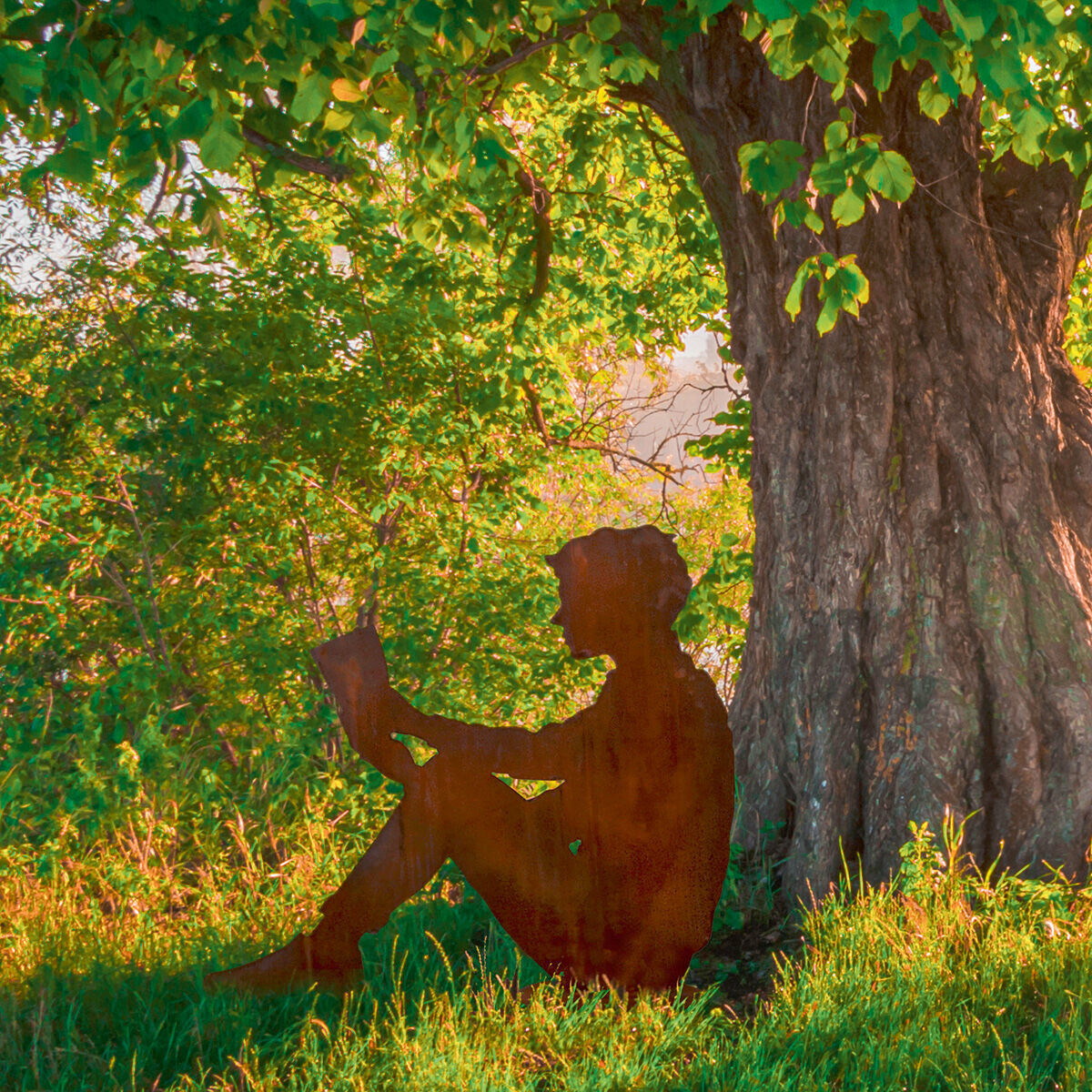 Eine Gartenfigur in Silhouette eines lesenden Menschen aus Metall sitzt neben einem großen Baum im Grünen. Die Figur hat eine Rost-Optik und befindet sich in einer naturreichen Gartenszene.