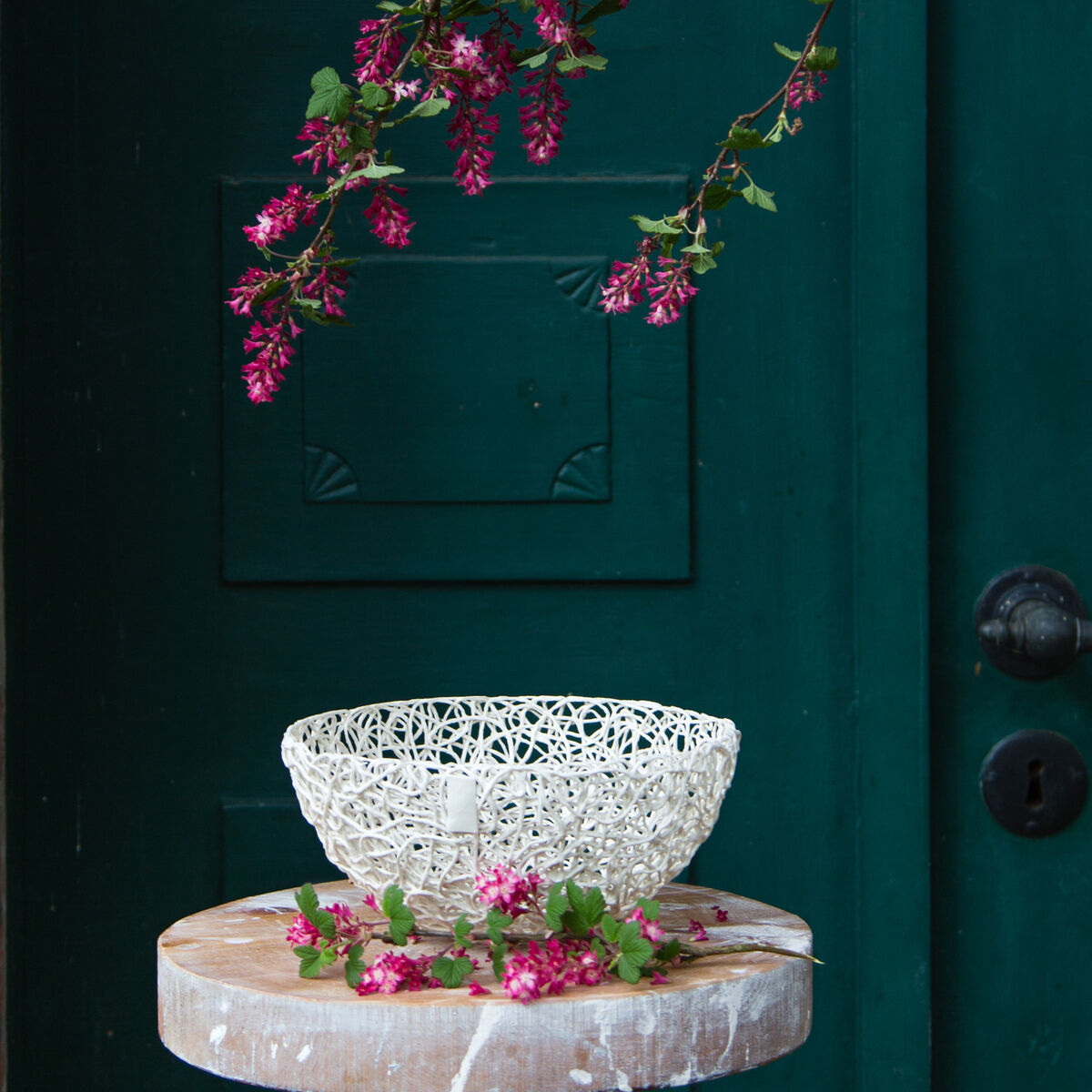 Eine weiße Porzellanschale steht auf einem runden, hölzernen Hocker. Der Hintergrund ist eine türkisfarbene Tür mit rosa Blumenranken, die von oben herabhängen.