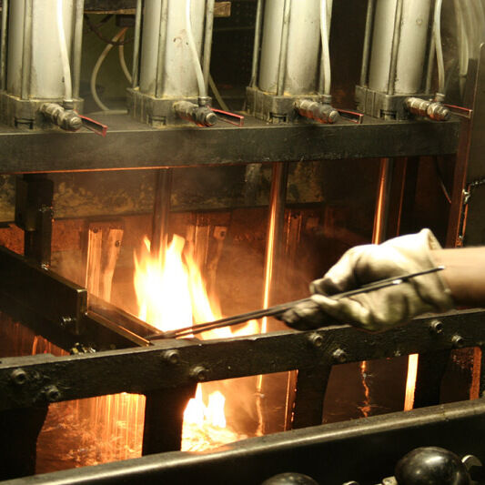 Ein Chinesisches Kochmesser, das in einem industriellen Verfahren über Flammen gehärtet wird. Ein Arbeiter mit Handschuh hält das Messer in die heiße Flamme. Mehrere Metallstangen und Maschinen sind ebenfalls sichtbar.