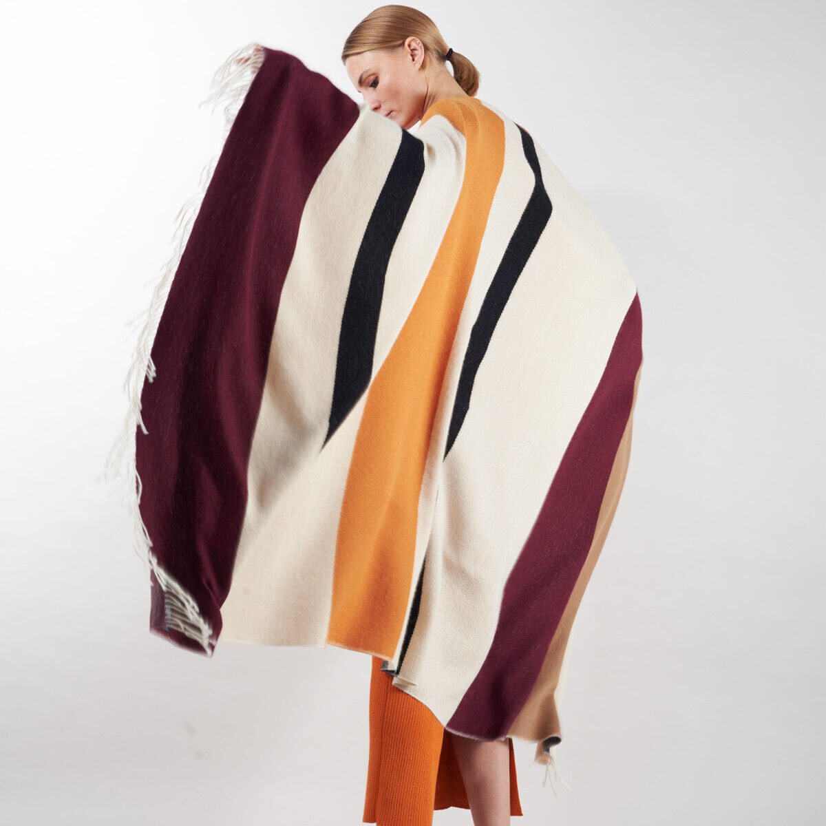 Eine Frau trägt ein elegantes Cape aus Alpaka-Wolle mit Streifen in den Farben Weiß, Schwarz, Orange und Bordeaux. Das Cape ist groß und warm, ideal für Herbst und Winter.