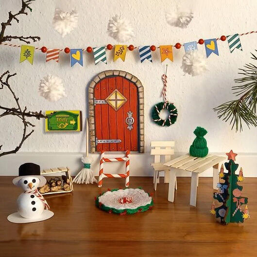 Der Adventskalender »Herzlich Willkommen, kleiner Wichtel.« von TOPP zeigt eine festlich dekorierte Szene mit kleiner Wichteltür, Miniaturmöbeln, Bastelarbeiten wie Schneemann und Weihnachtsbaum, einer Girlande, Zweigen und Watte-Schneeflocken vor weißer Wand.
