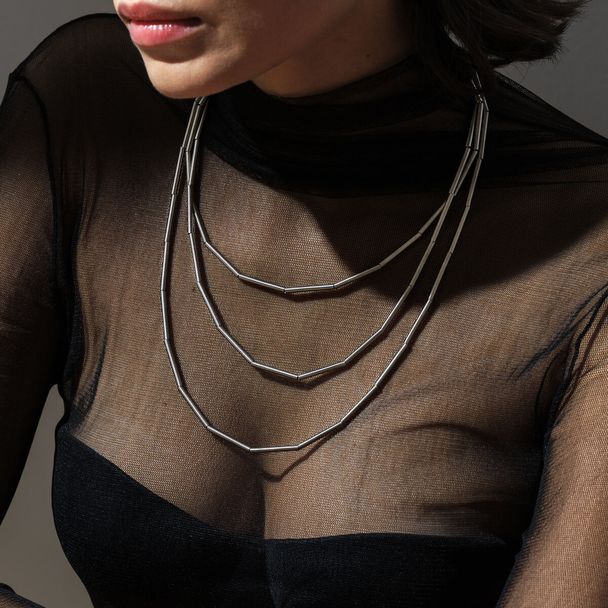 Eine Frau trägt das Collier "MISTY" von la molla aus Edelstahl, bestehend aus drei feinen, silbrig glänzenden Strängen. Die Frau hat einen schwarzen, halbtransparenten Rollkragen und sitzt in leichtem Schatten. Das Collier wirkt elegant und modern.