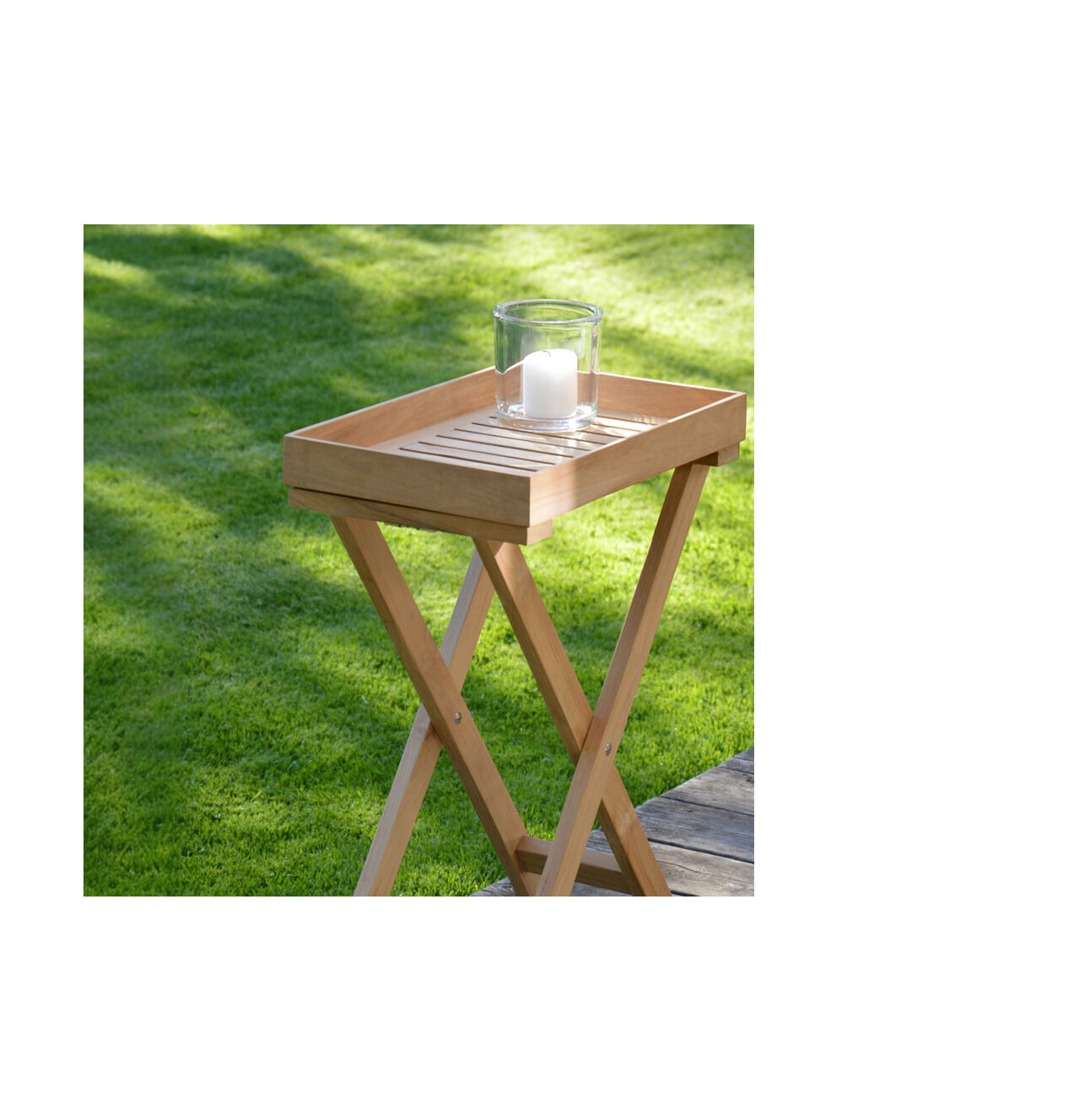 Ein schmaler, klappbarer Beistelltisch aus recyceltem Teakholz steht auf einer Terrasse neben einer Rasenfläche. Auf der abnehmbaren Tablettfläche steht ein Glas mit einer weißen Kerze. Das Holz zeigt natürliche Maserungen und Astlöcher.