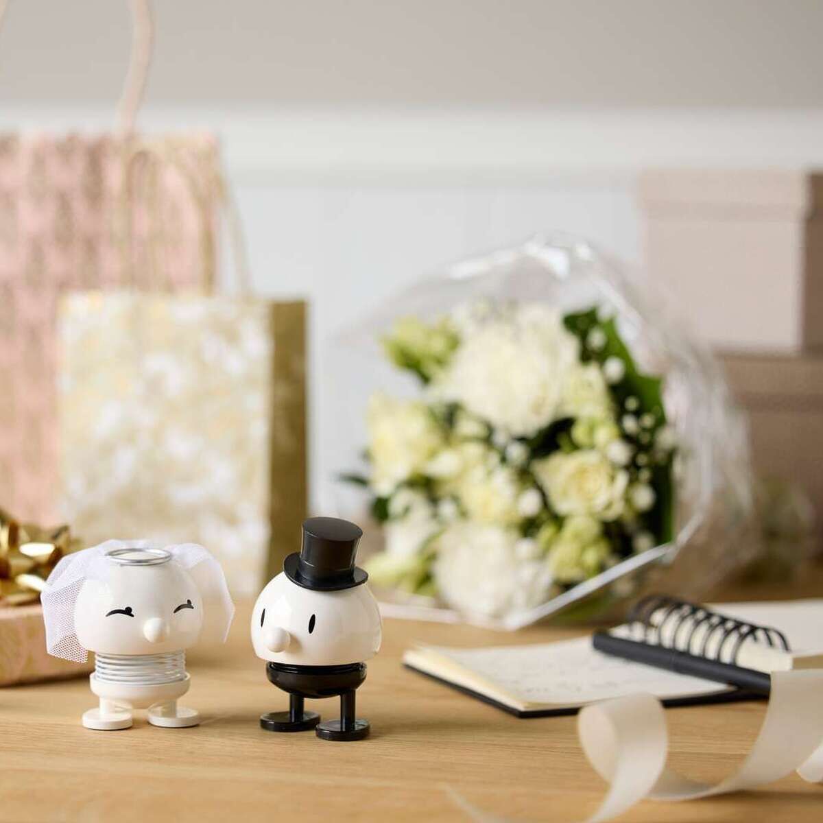 Ein dekorativer Tisch mit zwei Hoptimist-Figuren als Braut und Bräutigam aus ABS/Stahl, umgeben von weißen Blumen, Geschenkboxen, Notizbuch, goldenem Geschenkband und Sektgläsern. Im Hintergrund eine hell gemusterte Geschenktüte und ein Blumenstrauß.