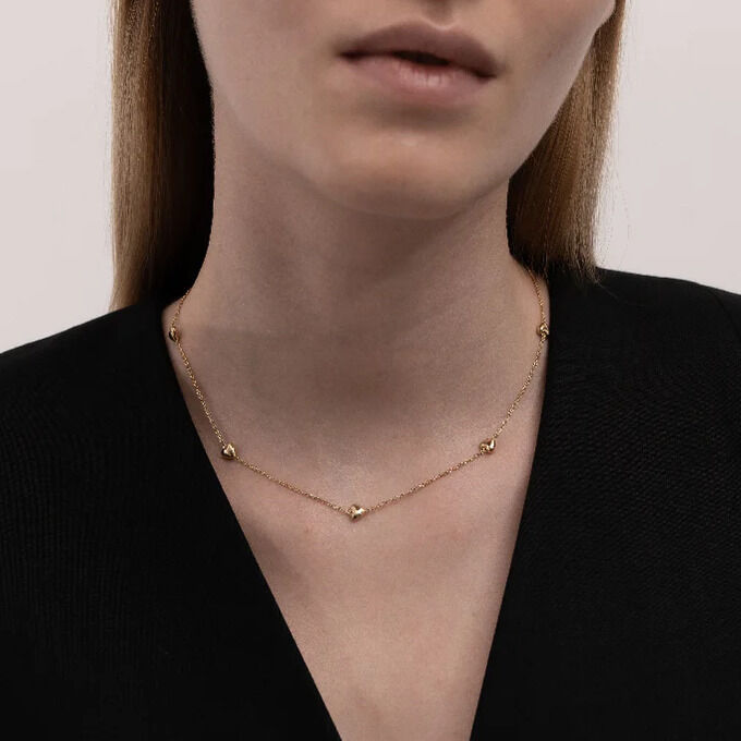 Eine Frau mit glattem, hellbraunem Haar trägt ein feines, vergoldetes Collier mit kleinen Herzanhängern von SKULTUNA MESSINGSBRUK. Sie trägt ein elegantes schwarzes Oberteil. Der Hintergrund ist hell und schlicht, was den Fokus auf das dezente Collier legt.