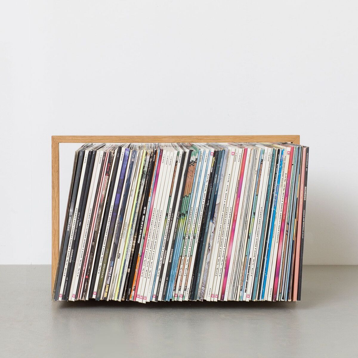 Ein Plattenständer aus Holz, der eine Sammlung von Vinyl-Schallplatten hält. Der Ständer hat eine minimalistische und moderne Gestaltung und steht vor einer weißen Wand auf einer grauen Oberfläche.