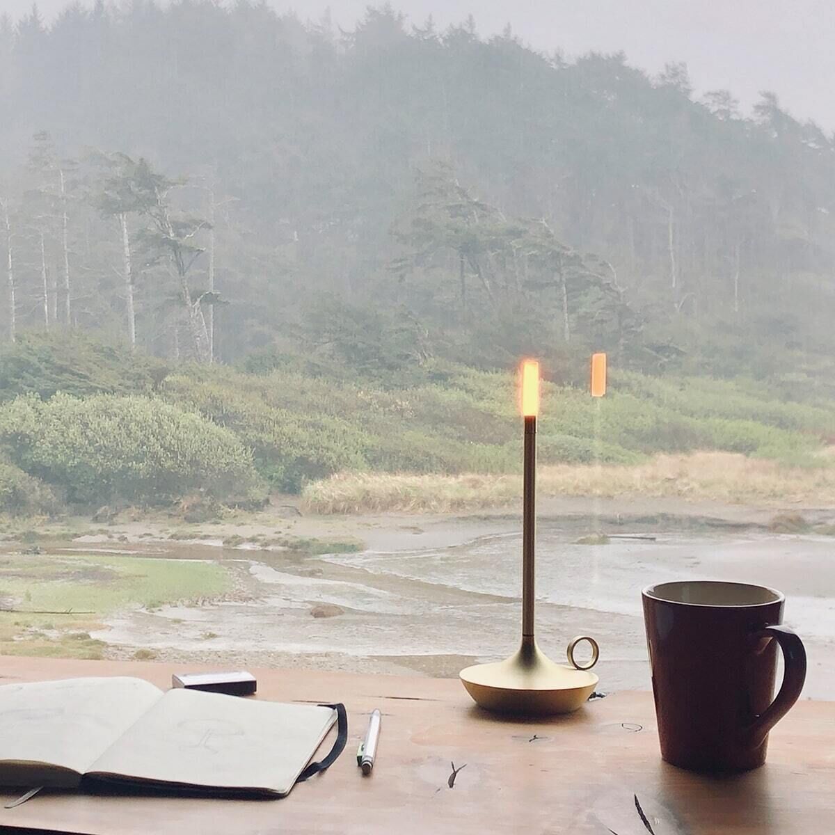 Eine stilvolle, goldfarbene Wick Tischlampe steht auf einem Tisch vor einem großen Fenster mit Blick auf eine nebelige Waldlandschaft. Auf dem Tisch befinden sich ein Notizbuch, ein Stift und eine braune Tasse. Die Lampe leuchtet und schafft eine gemütliche Atmosphäre.
