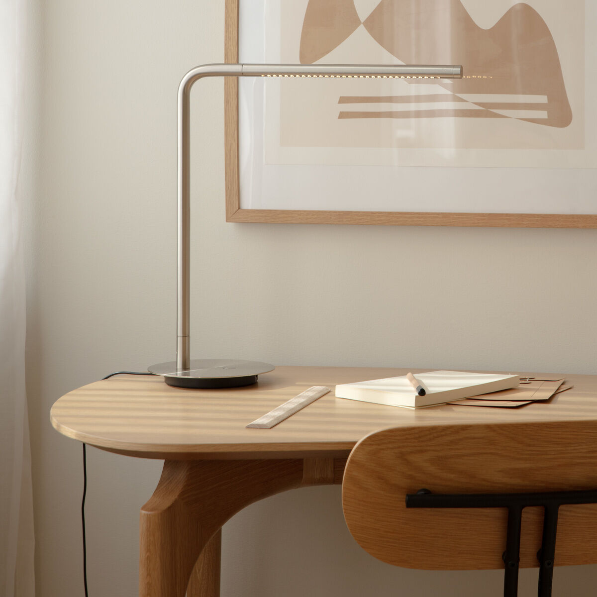 Eine minimalistische Tischleuchte 'Omni Table V2' steht auf einem hellen Holzschreibtisch vor einer modernen abstrakten Kunst an der Wand, neben einem Notizbuch, Lineal und Stift. Das Licht strahlt auf die Arbeitsfläche, die gesamte Szene wirkt hell und elegant.