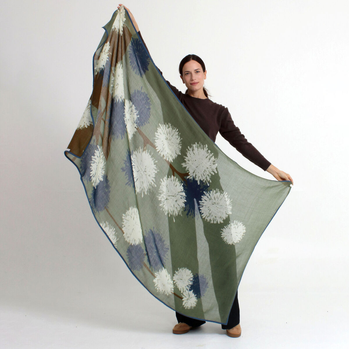 Frau hält einen großen, leichten Wollschal »Carol« von Shalls Barcelona hoch, sandfarben mit weißen und bläulichen floralen Mustern, ohne Fransen, dezente Umrandung. Schal aus 100% Schurwolle, modern und elegant inszeniert vor neutralem Hintergrund.