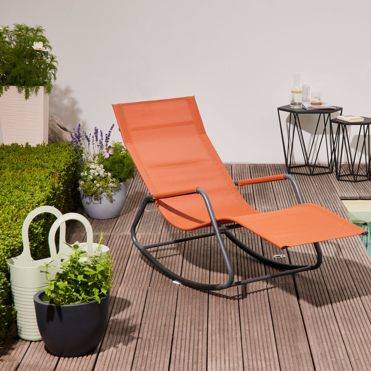 Moderne orangefarbene Swingliege »Aliano« mit Metallgestell steht auf Holzterrasse neben einem Pool, umgeben von Pflanzen, Gießkanne und kleinen Tischen, bei sonnigem Wetter. Outdoor-Szenerie lädt zur Entspannung ein.