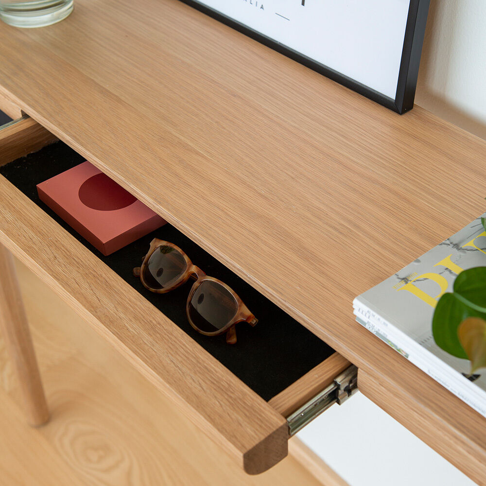 Detailaufnahme des Konsolentischs »Heart'n'Soul« von Umage aus massiver Eiche. Die geöffnete, schwarze Stoffschublade enthält eine braune Sonnenbrille und eine rosafarbene Schachtel. Auf der Tischplatte liegen ein Magazin, eine Pflanze und ein gerahmtes Bild.