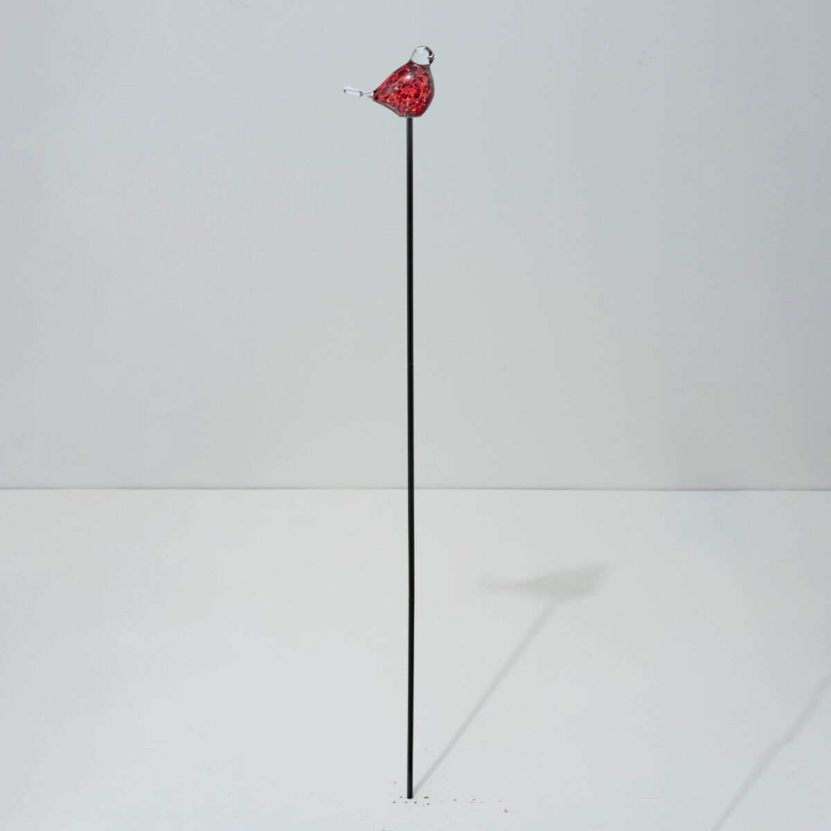 Glas-Gartenstecker von Ars mundi: Ein kleiner roter Vogel aus Glas sitzt auf einem langen, dünnen, schwarzen Metallstab vor neutralem Hintergrund. Die Skulptur ist minimalistisch gehalten und dient als dekoratives Gartenelement.