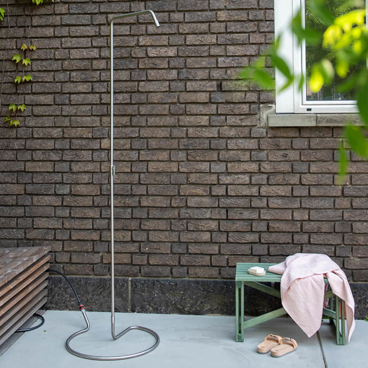 Gartendusche »Serpentine« neben einer Ziegelsteinmauer, mit einem kleinen, grünen Tisch, einem Handtuch und schlappen im minimalistischen Garten.