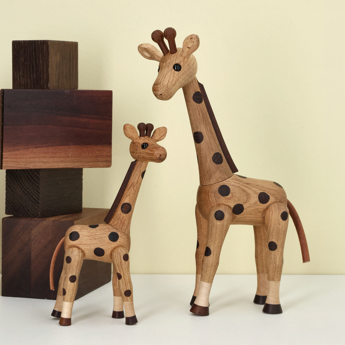 Zwei hölzerne Giraffenfiguren, eine groß und eine klein, mit dunklen Punkten stehen auf einer weißen Oberfläche vor hellgelbem Hintergrund. Links hinten sind dekorative Holzwürfel in verschiedenen Braunschattierungen gestapelt.