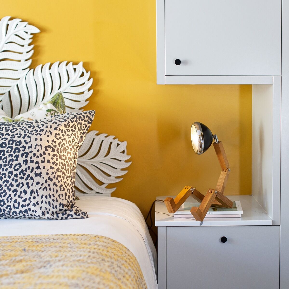 Eine stilvolle Tischleuchte namens »Mr. Wattson« steht auf einem Nachttisch neben einem Bett. Die Wand hinter dem Bett ist gelb gestrichen und das Kopfteil besteht aus weißen, blattähnlichen Strukturen. Neben der Lampe liegen einige Bücher.