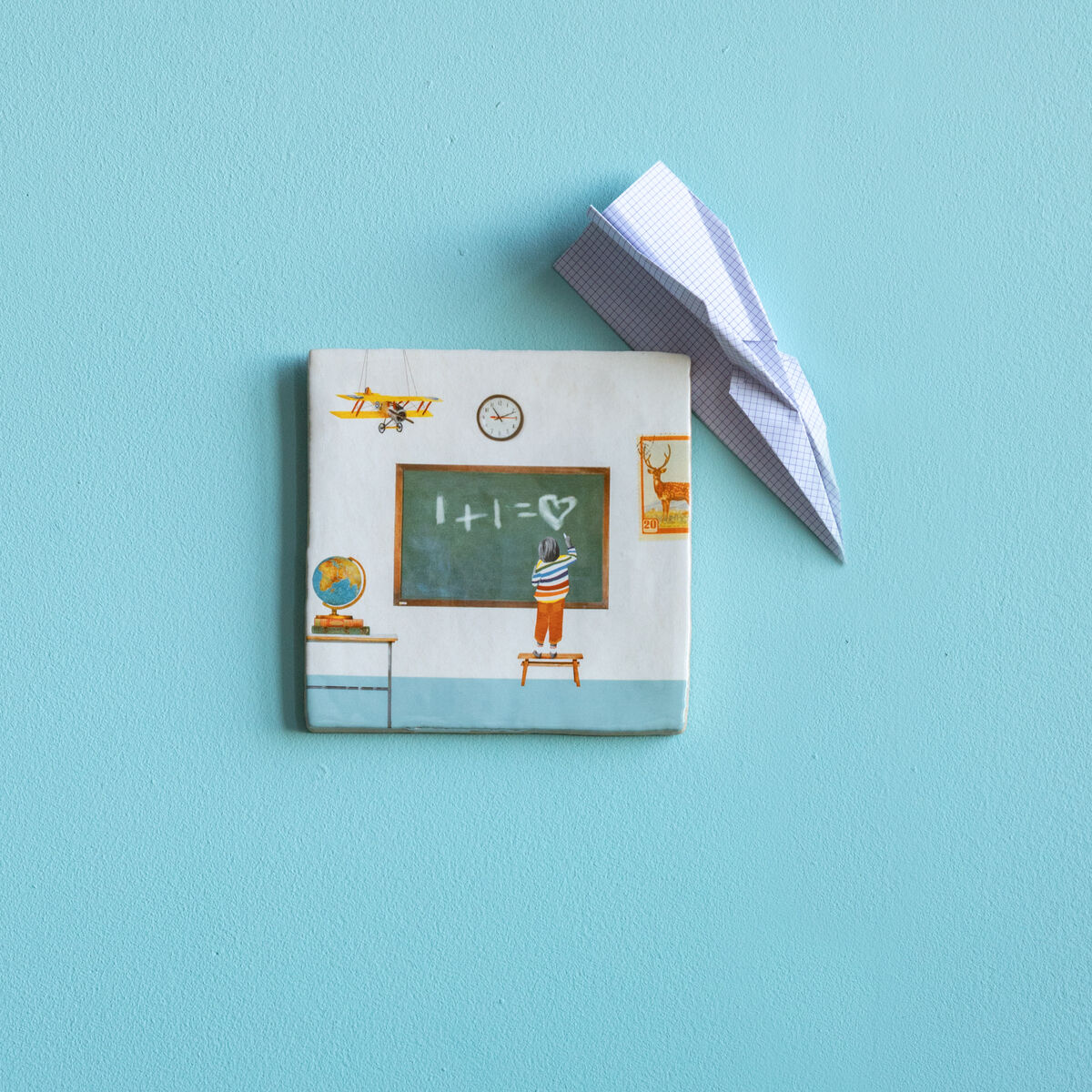 Eine Keramikfliese mit einer Zeichnung eines Klassenzimmers hängt an einer blauen Wand. Auf der Fliese sind eine Tafel mit einer Rechenaufgabe (1+1=2), ein Globus und Stühle abgebildet. Daneben sind Papierflieger. Unten ist ein Holztisch mit Büchern und einem Globus zu sehen.